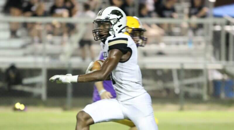 A football player in a white uniform carries the ball with a defender behind him.